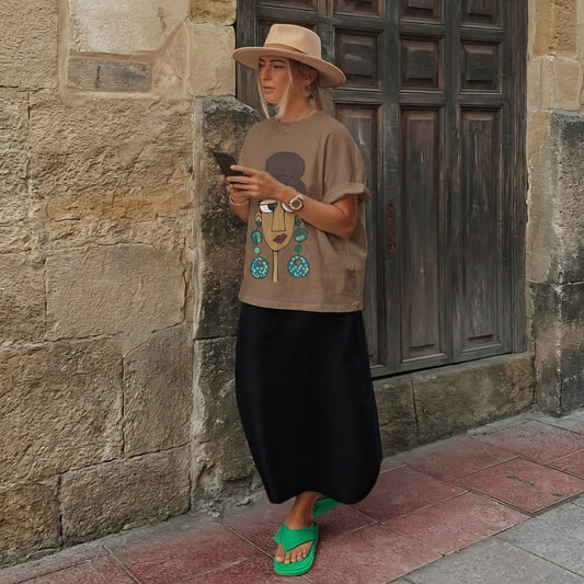 Woman wearing a brown t-shirt with a graphic design, black skirt, and green sandals standing against a stone wall.