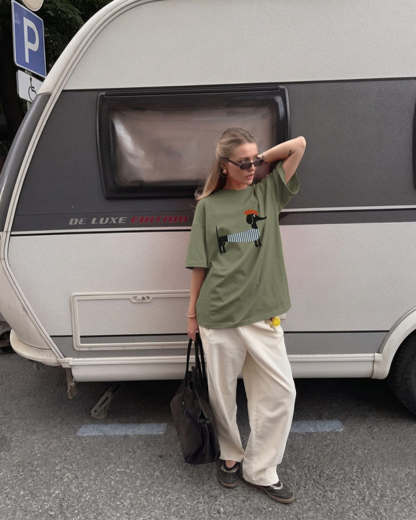 A woman with sleek blonde hair and sunglasses wears an oversized olive The Parisian Pup™ tee with cream corduroy trousers, leaning on a vintage caravan.