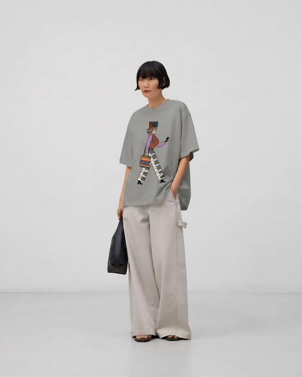 A woman with a black bob wears an oversized gray The Urban Rhythm™ tee with wide stone-colored trousers against a plain studio backdrop.