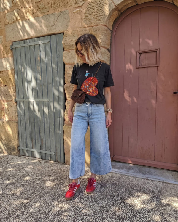 A woman with a wavy blonde bob and sunglasses wears The Heart Grower™ tee in black with light wide-leg jeans, a leopard crossbody bag and red sneakers, standing before sage and rose wooden doors.