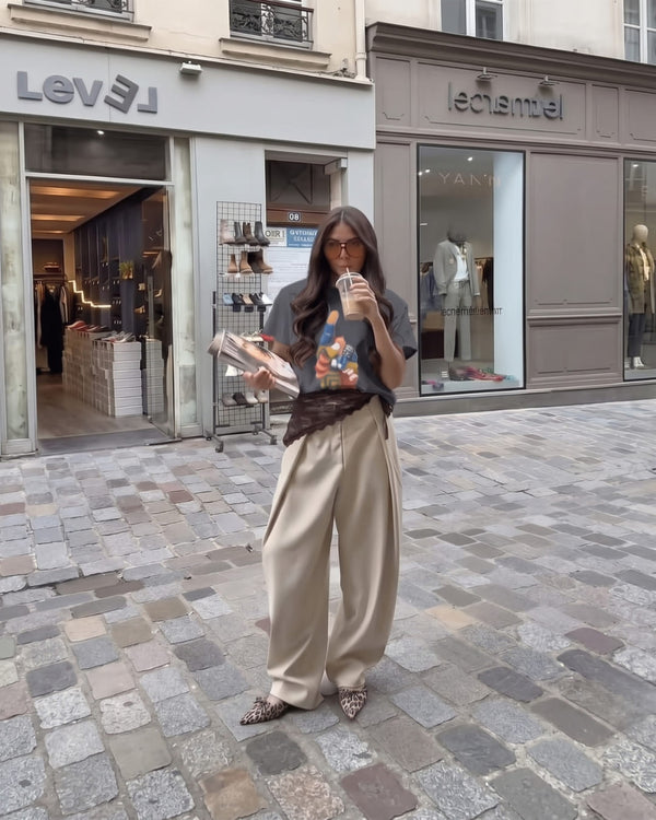 A woman with long dark hair sips an iced coffee in The Painted Luck™ tee in charcoal, beige wide-leg trousers and leopard flats on a cobblestone street.