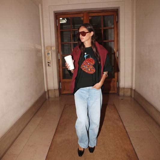 Person standing in a hallway holding a cup, wearing a black t-shirt with a colorful design and light blue jeans.