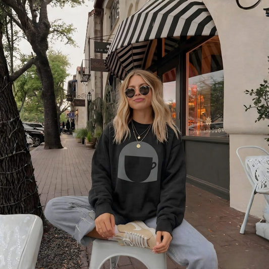 Woman sitting on a stool in an urban setting wearing a black sweatshirt with a coffee cup graphic.