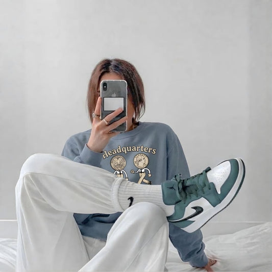 Person wearing a blue sweatshirt and white pants sitting on a bed, taking a mirror selfie.