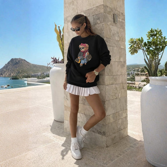Woman wearing a black sweatshirt with a graphic design, white skirt, and white sneakers, standing outdoors by a stone column with scenic background.
