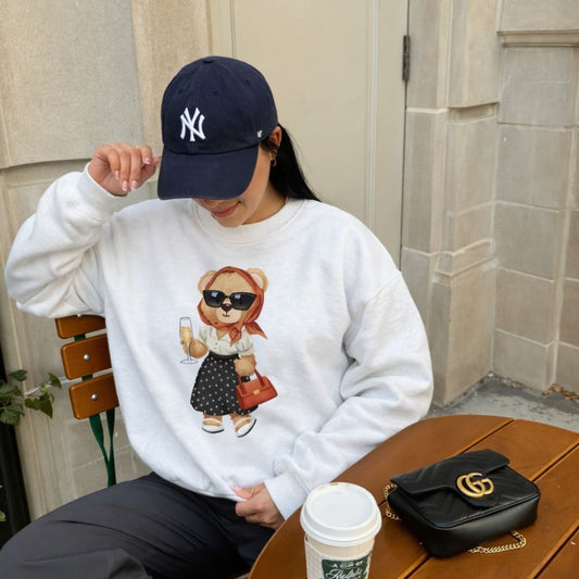 Person wearing a white sweatshirt with a bear graphic, sitting outdoors with a coffee cup and black handbag.