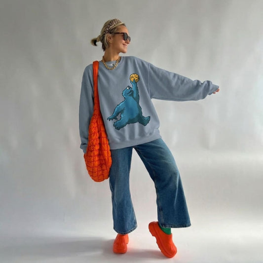 Person wearing a light blue sweatshirt with a bear graphic, blue jeans, and orange shoes, holding an orange bag.