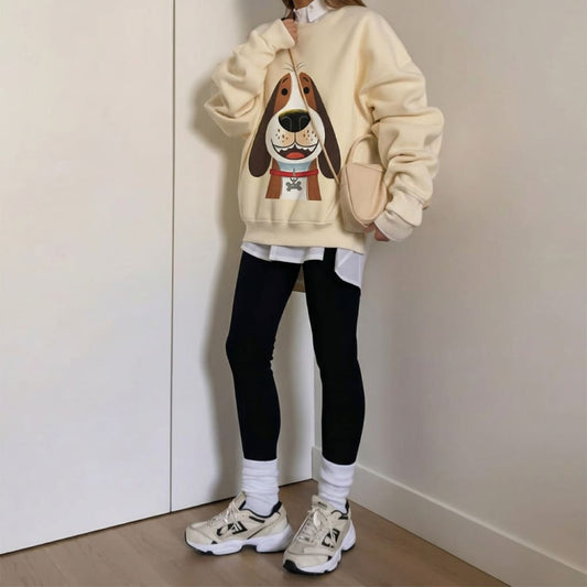 Person wearing a beige sweatshirt with a dog graphic, black leggings, and white sneakers in a minimal indoor setting.