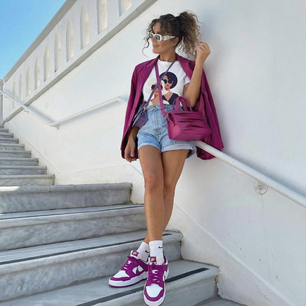 Woman in stylish outfit with purple jacket, shorts, and sneakers standing on stairs.