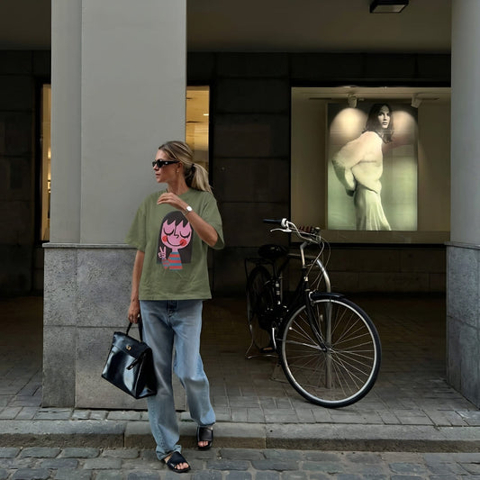 Woman in a green t-shirt with a graphic design, standing next to a bicycle on a city street.