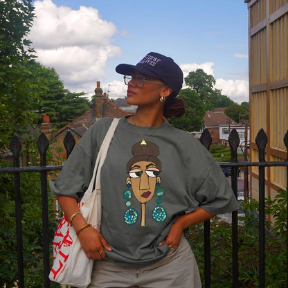 Person wearing a gray t-shirt with a graphic design, standing outdoors with a black metal fence and greenery in the background.