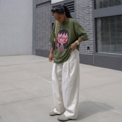 Person wearing a green oversized shirt with a graphic design and white pants standing on a sidewalk.