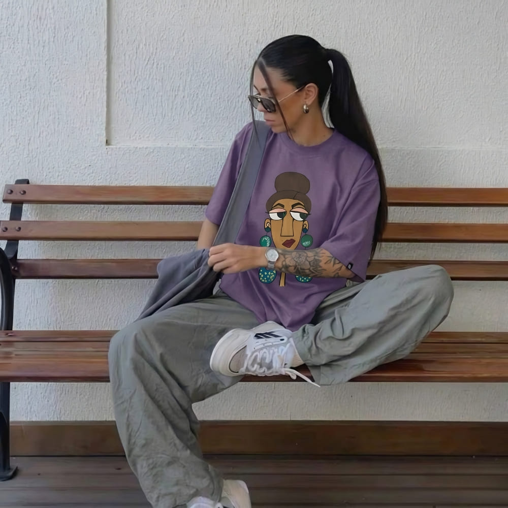 Person sitting on a bench wearing a grape purple t-shirt with a graphic design, gray pants, and white sneakers.