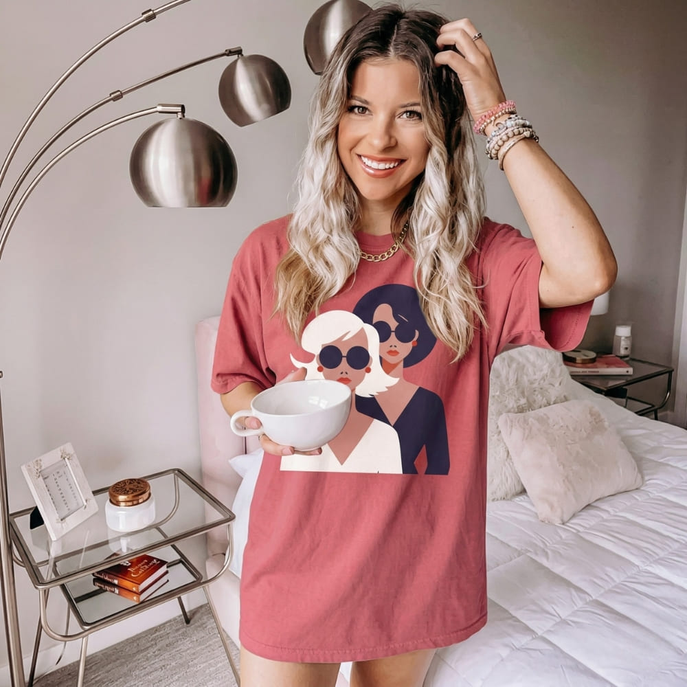 Woman wearing a pink t-shirt with graphic design, holding a mug in a bedroom setting.