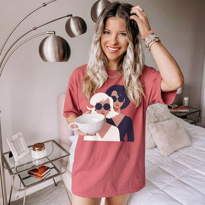 Woman wearing a pink t-shirt with graphic design, holding a mug in a bedroom setting.
