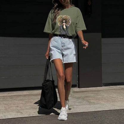 Person wearing a green t-shirt with a graphic design, light blue denim shorts, and white sneakers, standing against a dark wall.