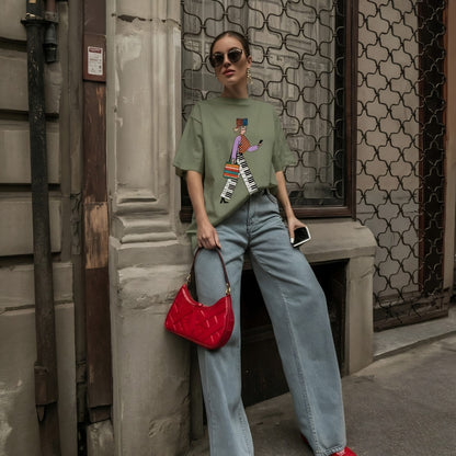 Woman wearing a green t-shirt with a colorful design, blue jeans, and red shoes, holding a red handbag, standing against a textured wall.