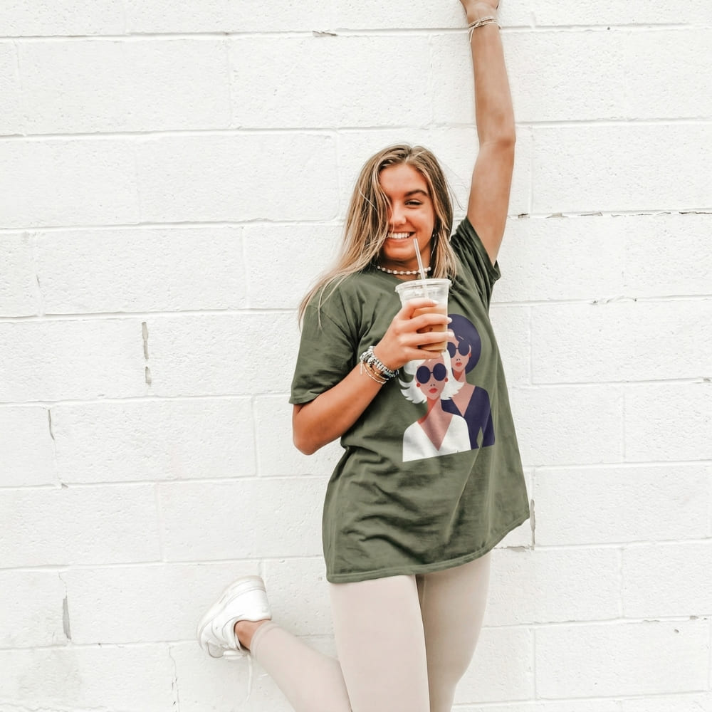 Woman wearing a green t-shirt with a graphic design, holding a cup against a white brick wall.