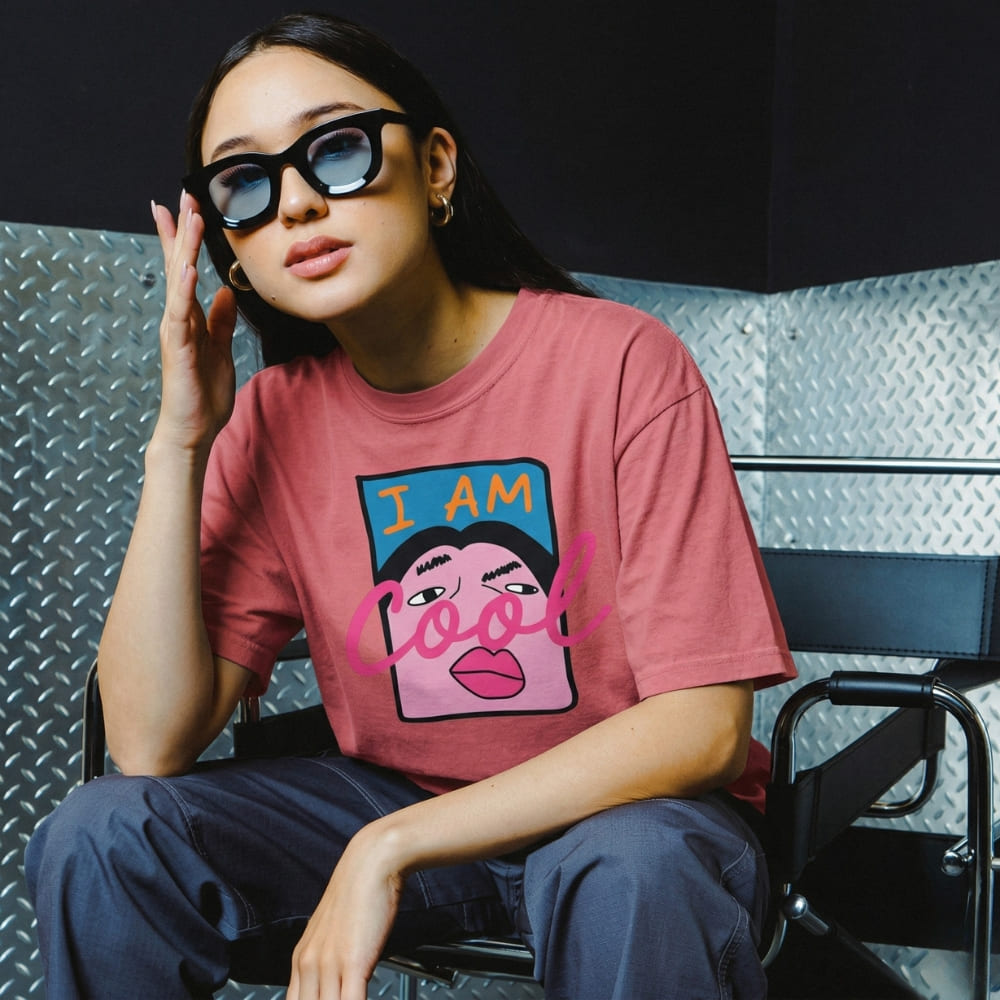 Person wearing a pink t-shirt with a graphic design sitting against a metallic wall.