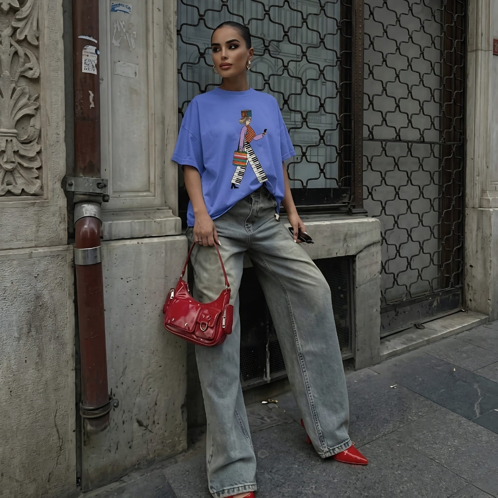 Person wearing a blue t-shirt with a colorful design, light blue jeans, and red shoes, holding a red handbag against a textured wall.