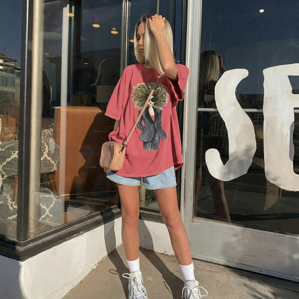 Woman wearing a pink oversized t-shirt with a graphic design, denim shorts, and white sneakers, standing in front of a store window.