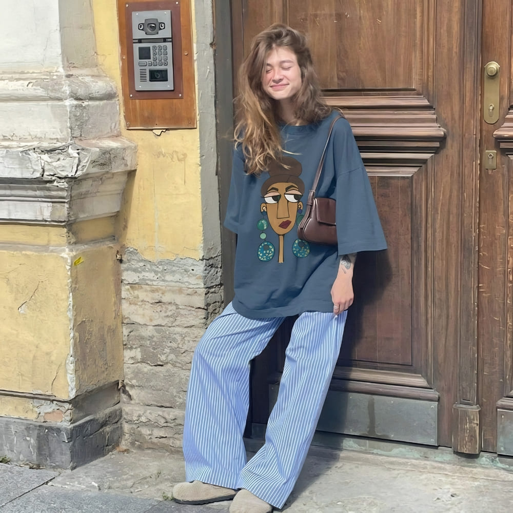 Woman wearing a mid-night blue oversized shirt with a graphic design, standing against a textured wall and wooden door.