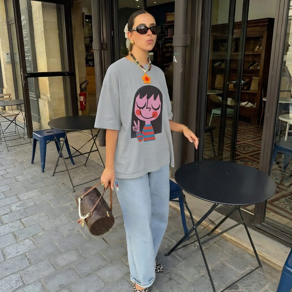 Woman wearing a graphic t-shirt and sunglasses, standing outside a building with chairs and tables.