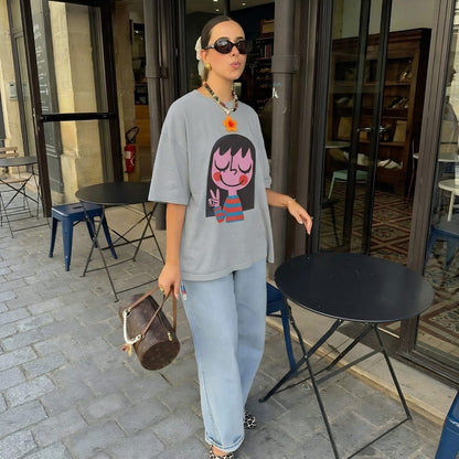 Woman wearing a graphic t-shirt and sunglasses, standing outside a building with chairs and tables.