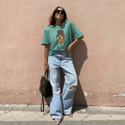 Woman wearing a teal t-shirt with a graphic design and light blue jeans against a pink wall.