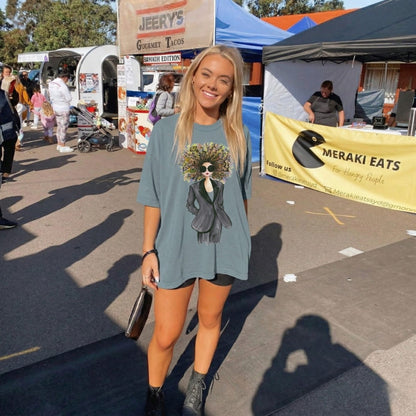 Woman in a ice-blue t-shirt with a graphic design at an outdoor event with tents and people.