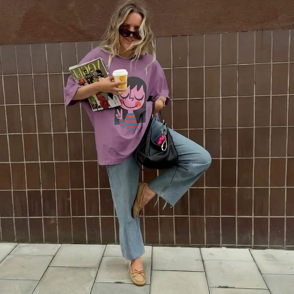 Person in a purple shirt with a character design, holding books and a coffee cup, standing against a tiled wall.