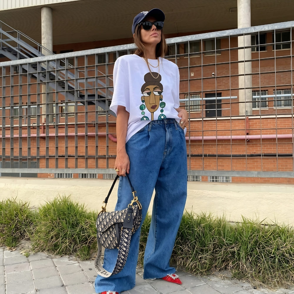 Woman wearing a white t-shirt with a graphic design, blue jeans, and a cap, standing outdoors.