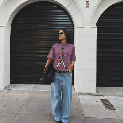 Woman wearing a purple graphic t-shirt and blue jeans standing in front of a building with black shutters.
