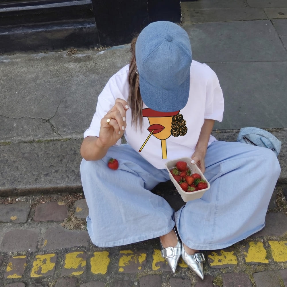 Person sitting on a step with a box of strawberries and wearing a white t-shirt with a colorful design and light blue jeans.