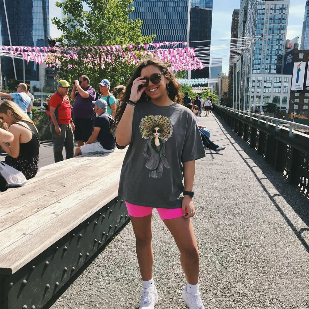Woman in a pepper-gray t-shirt and pink shorts standing on a city street with people and buildings in the background.