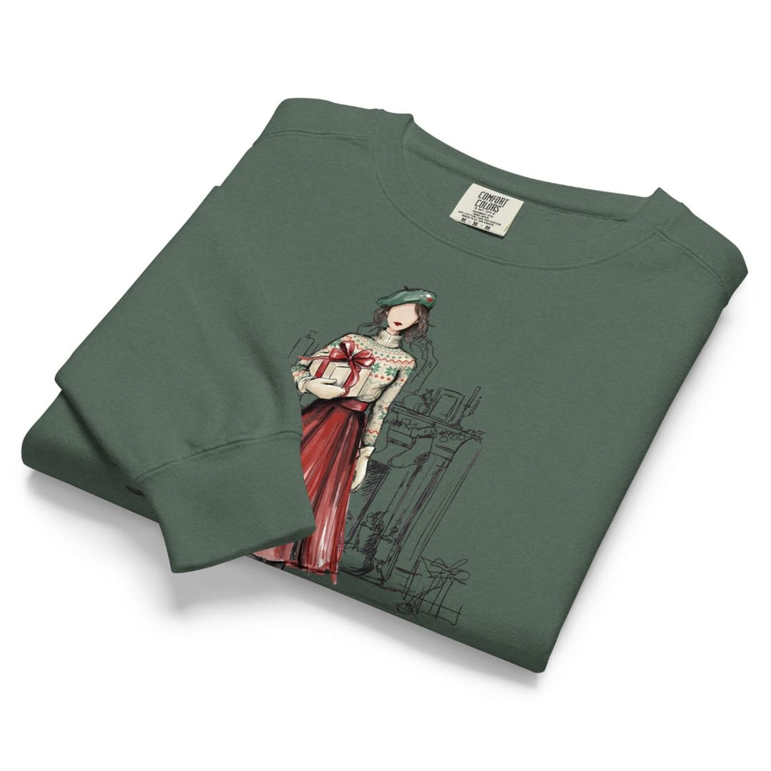 Holiday Muse™ sweatshirt by Blossombless.co in Blue Spruce, neatly folded to show the garment-dyed fleece and a sketch-style watercolor illustration of a faceless woman in a green beret, Fair Isle sweater and red pleated skirt holding a wrapped gift