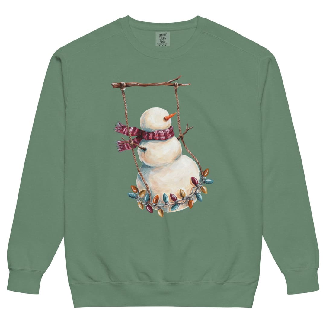 Swing Season™ sweatshirt by Blossombless.co in Light Green, front view: a painterly snowman swinging on a strand of vintage Christmas lights from a bare winter branch, wearing a striped burgundy scarf