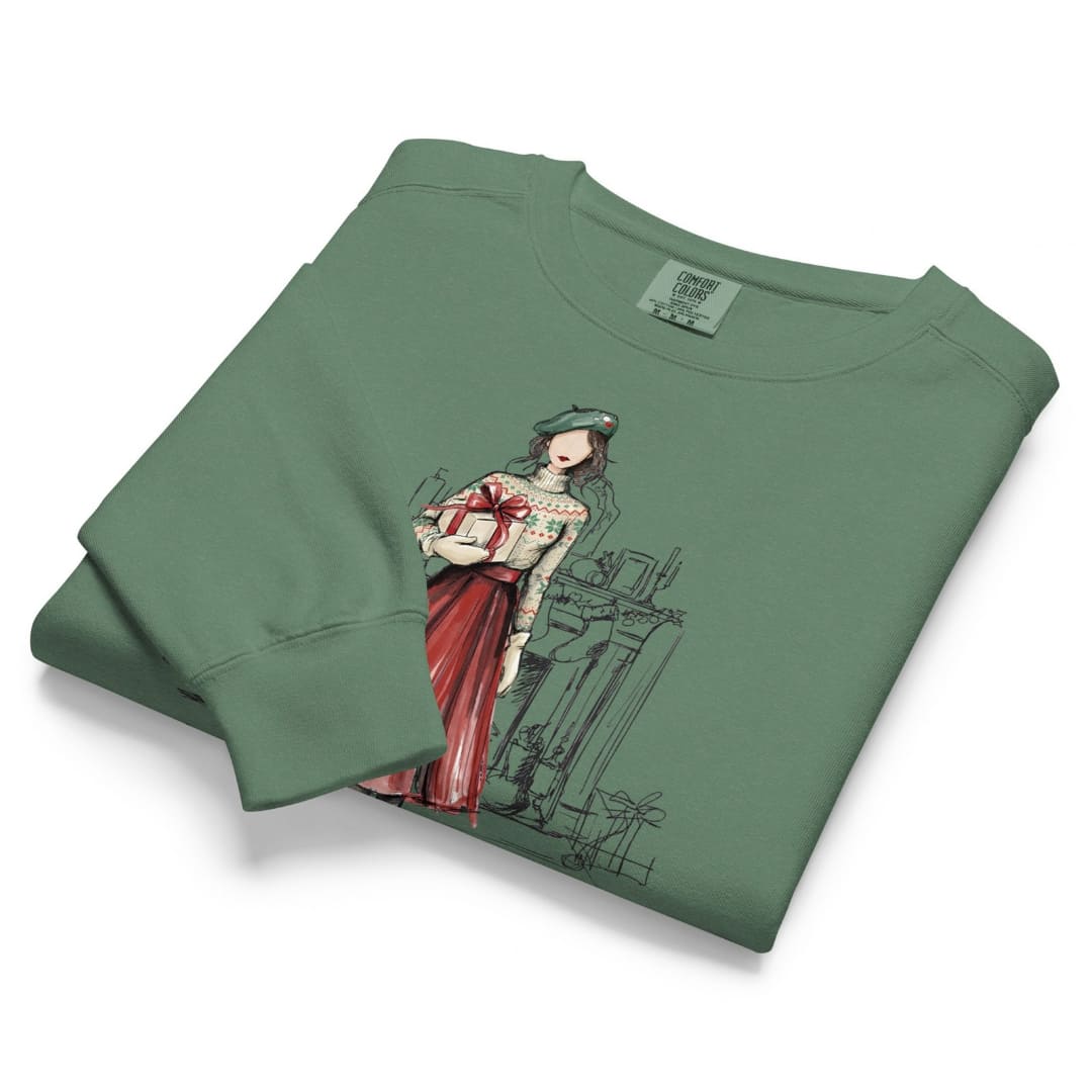 Holiday Muse™ sweatshirt by Blossombless.co in Light Green, neatly folded to show the garment-dyed fleece and a sketch-style watercolor illustration of a faceless woman in a green beret, Fair Isle sweater and red pleated skirt holding a wrapped gift