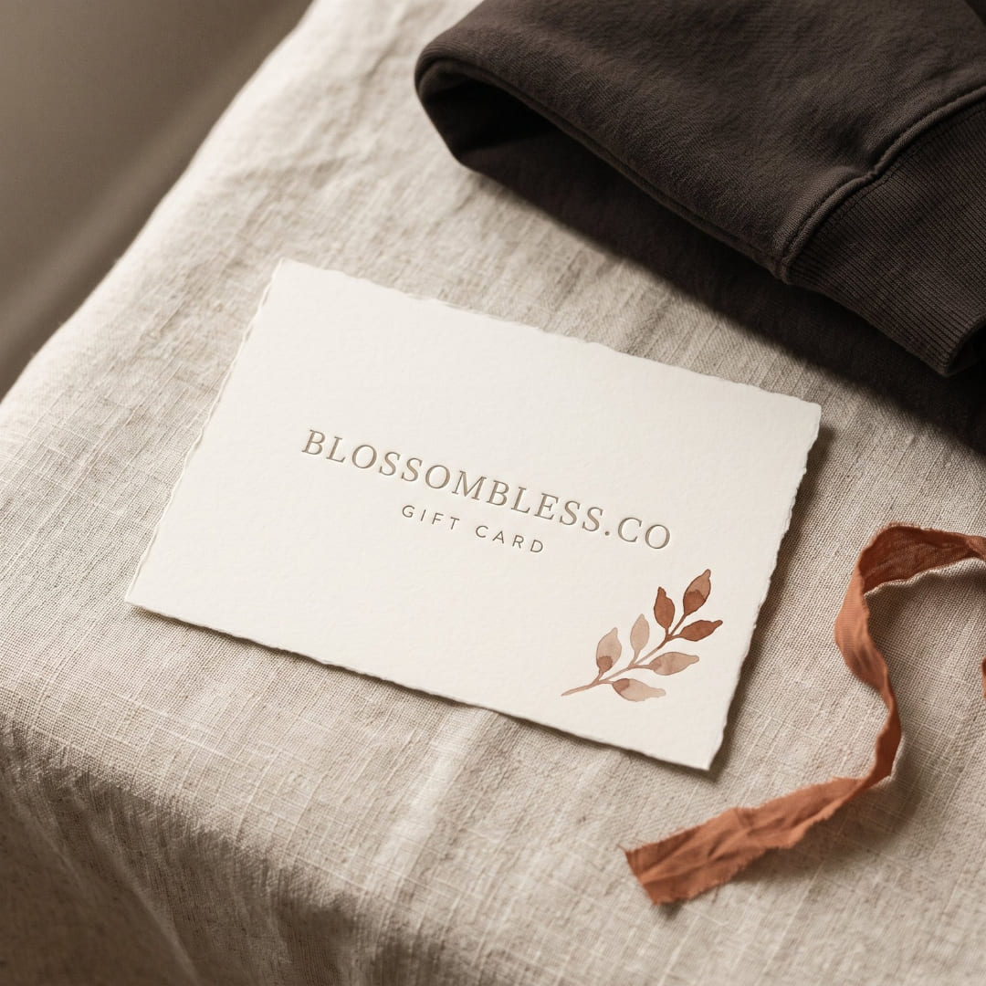Ivory Blossombless.co gift card with a hand-painted rust watercolor sprig, resting on natural linen beside a folded espresso brown sweatshirt and a rust silk ribbon