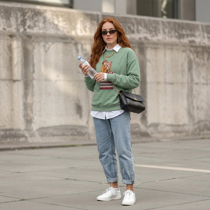 Woman wearing the Between Chapters™ sweatshirt by Blossombless.co in Light Green with sunglasses and light-wash jeans, standing by a stone wall outdoors