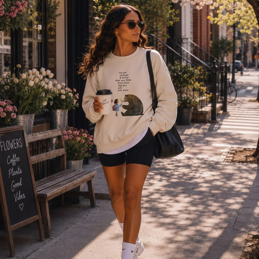 Stronger & Braver™ oversized sweatshirt by Blossombless.co in Dust, lifestyle: woman in sunglasses on a tree-lined brick sidewalk outside a café