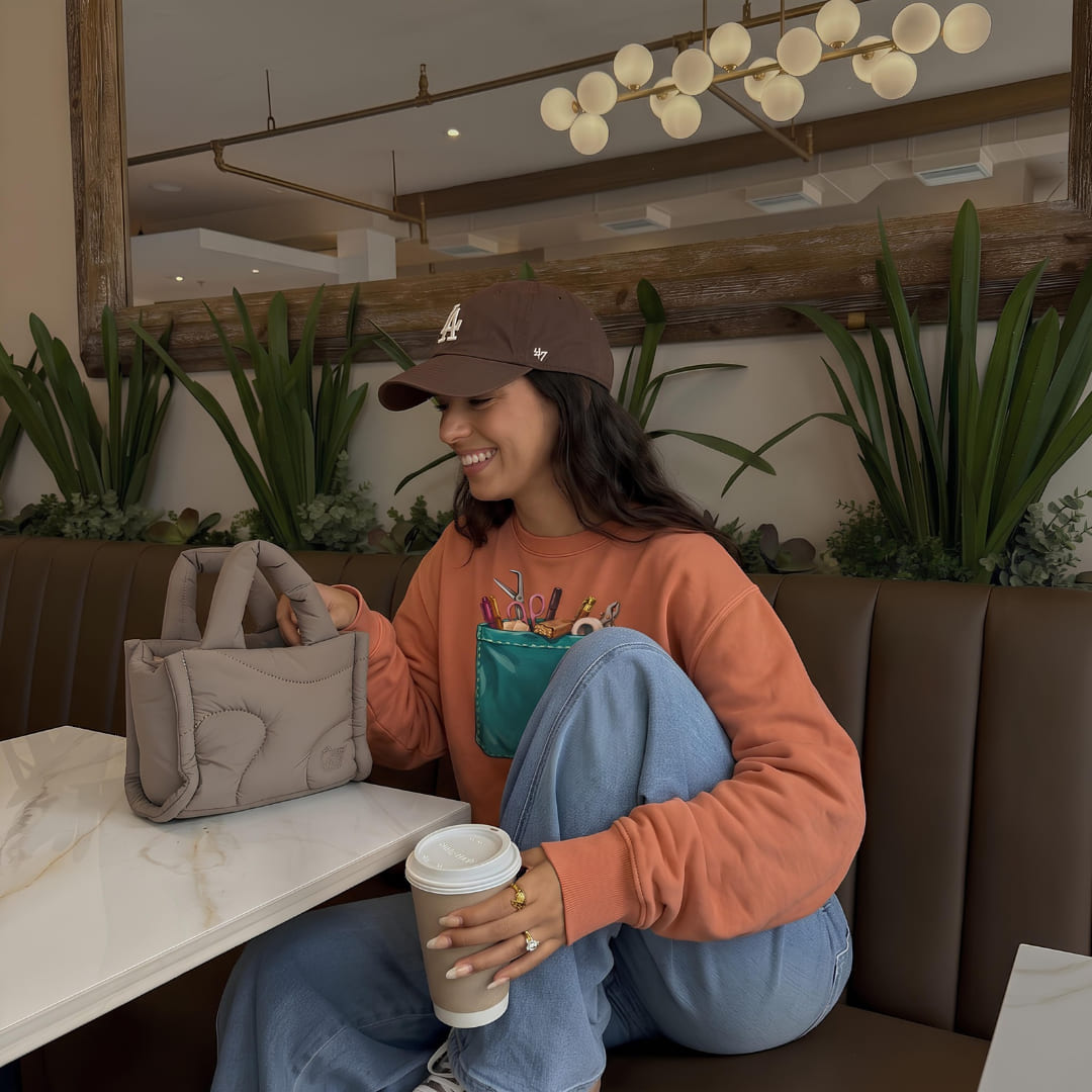 Woman wearing The Shift Pocket™ sweatshirt in Yam, sitting in a café with a coffee and a tote bag
