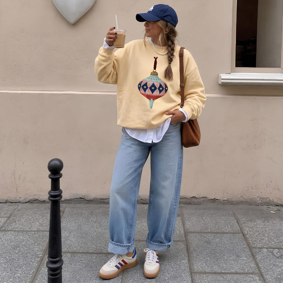 Woman wearing the Handed Down™ sweatshirt by Blossombless.co in Butter with a navy cap, light-wash jeans and a brown shoulder bag, sipping an iced coffee on a city sidewalk