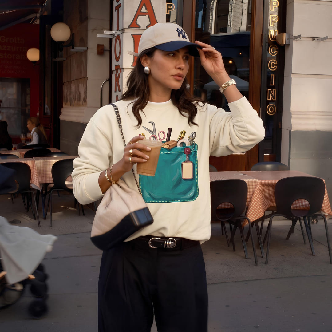 Woman wearing The Shift Pocket™ sweatshirt in Ivory outside a café, holding a coffee and wearing a cap