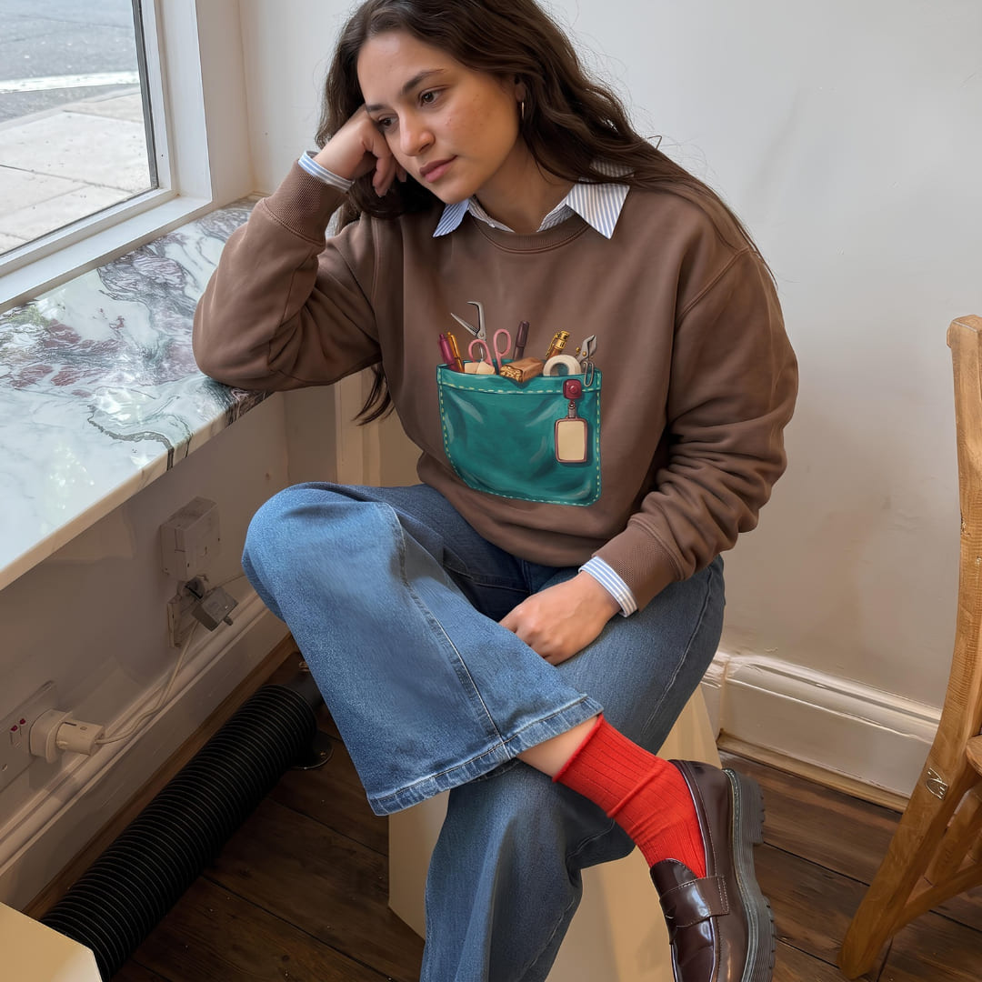 Woman wearing The Shift Pocket™ sweatshirt in Espresso, sitting by a window with jeans and red socks
