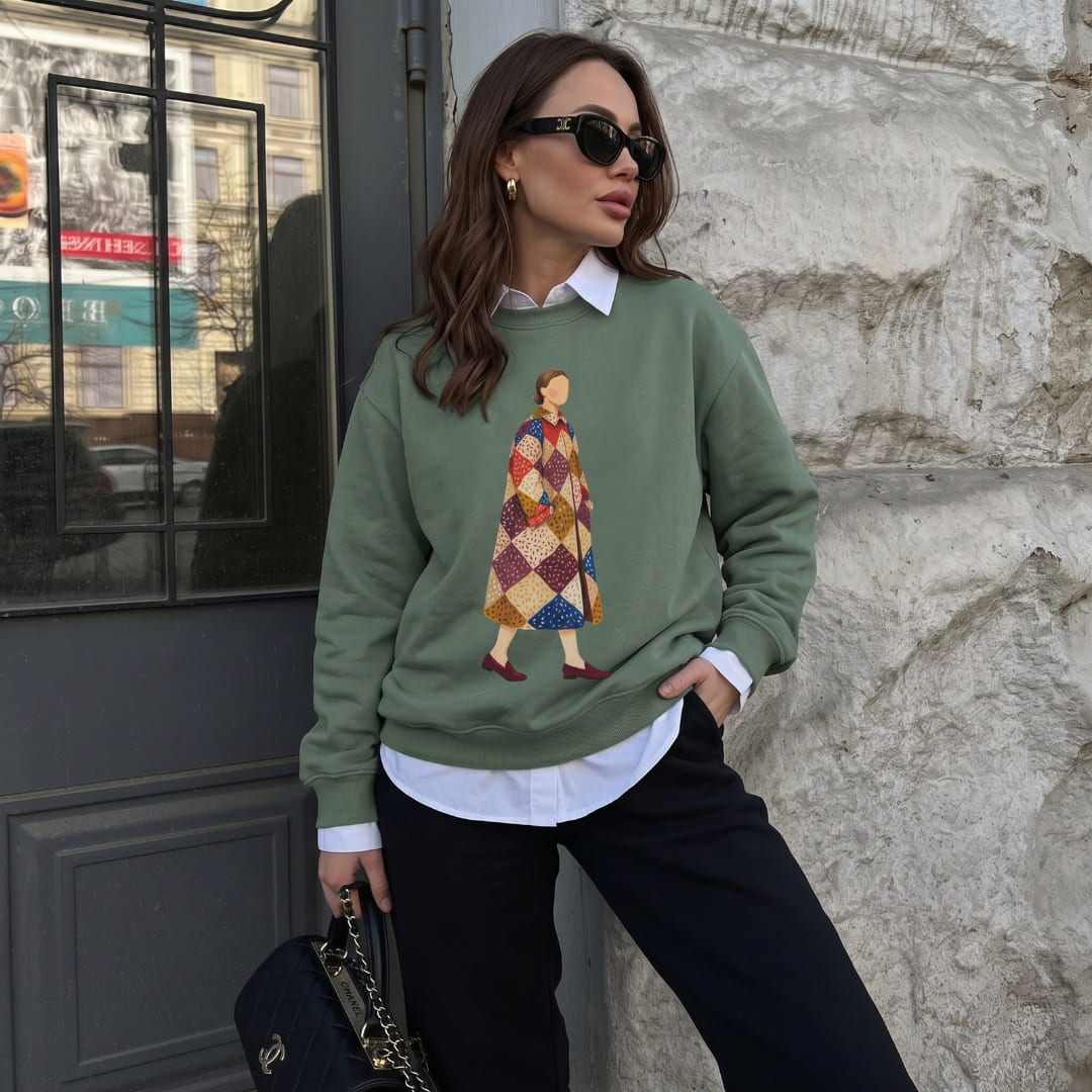 Woman wearing the Pieced Together™ sweatshirt by Blossombless.co in Blue Spruce over a white collared shirt with black trousers and sunglasses, leaning against a stone wall