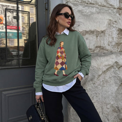 Woman wearing the Pieced Together™ sweatshirt by Blossombless.co in Blue Spruce over a white collared shirt with black trousers and sunglasses, leaning against a stone wall