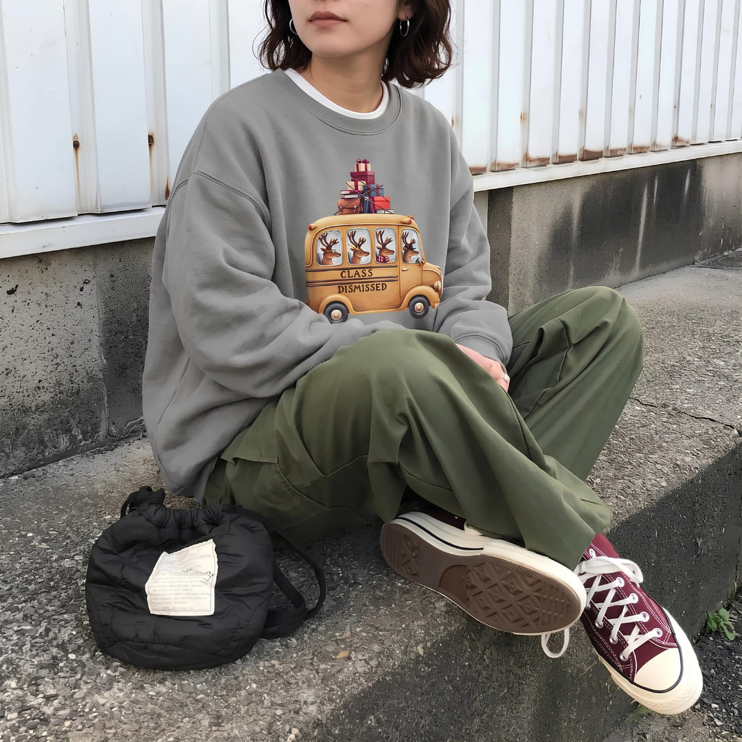 Woman wearing the Class Dismissed™ sweatshirt by Blossombless.co in Grey with olive trousers and burgundy sneakers, sitting on a curb beside a black backpack