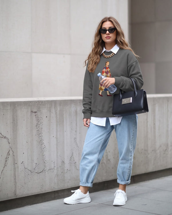 Woman wearing the Pieced Together™ sweatshirt by Blossombless.co in Pepper with light-wash jeans, a white collared shirt underneath, sunglasses and a black handbag, walking past a concrete wall