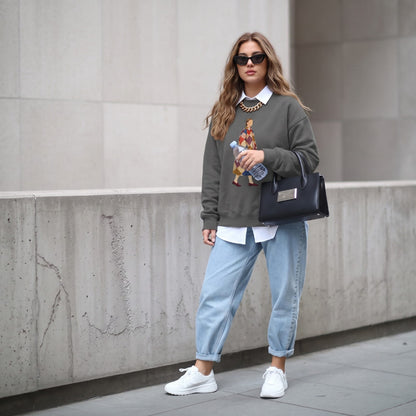 Woman wearing the Pieced Together™ sweatshirt by Blossombless.co in Pepper with light-wash jeans, a white collared shirt underneath, sunglasses and a black handbag, walking past a concrete wall